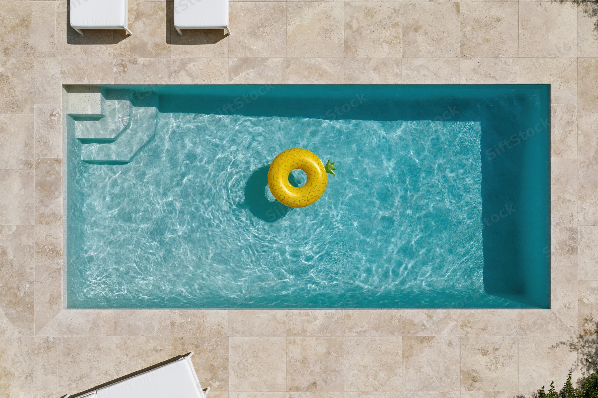 Aerial top-down view of a rectangular outdoor swimming pool with a yellow pineapple inflatable float in clear turquoise water surrounded by beige tiled patio and white sun loungers