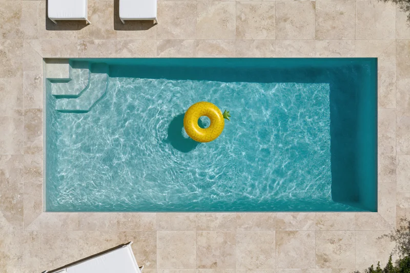 Aerial top-down view of a rectangular outdoor swimming pool with a yellow pineapple inflatable float in clear turquoise water surrounded by beige tiled patio and white sun loungers