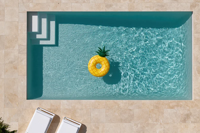 Aerial top-down view of a rectangular swimming pool with a pineapple float and lounge chairs on a tiled deck