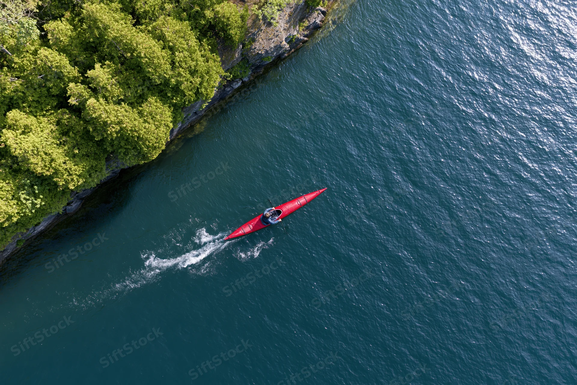 Person paddling a red kayak on a calm river near a forested shoreline