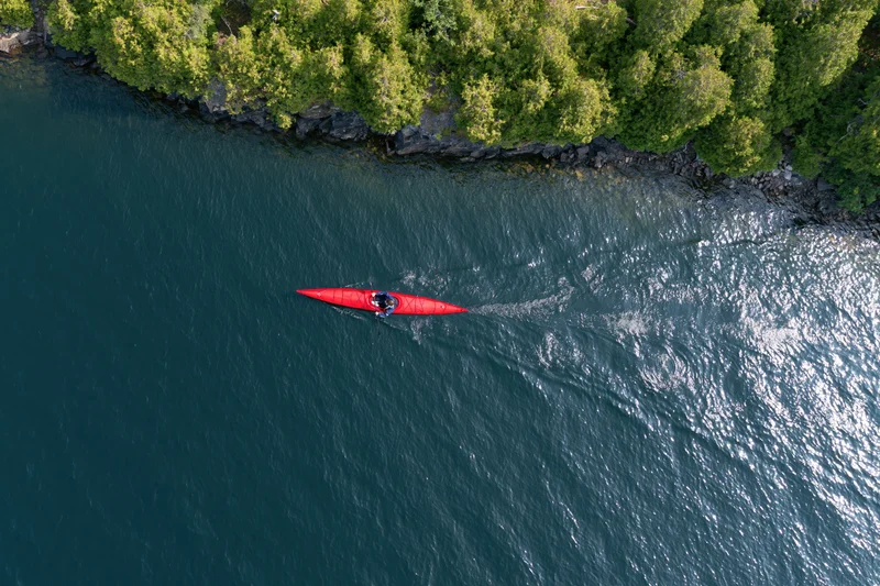 Aerial top-down view of a person paddling a red kayak on a calm river near a forested shoreline