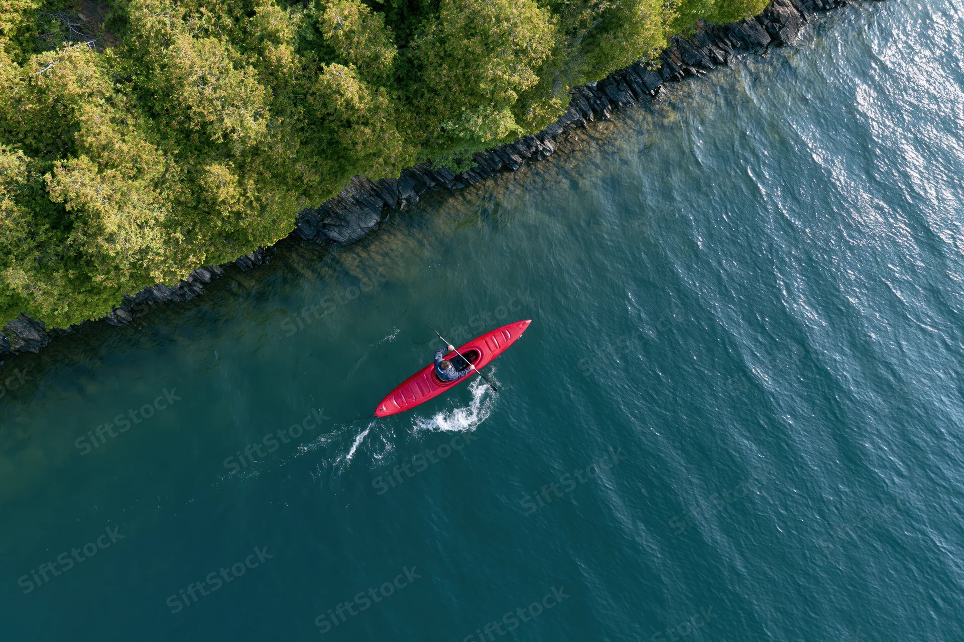 Person paddling red kayak on river near forested shoreline from aerial view