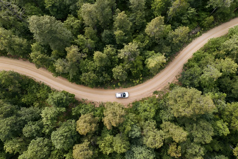 White car driving on winding dirt road through dense forest from aerial top-down view
