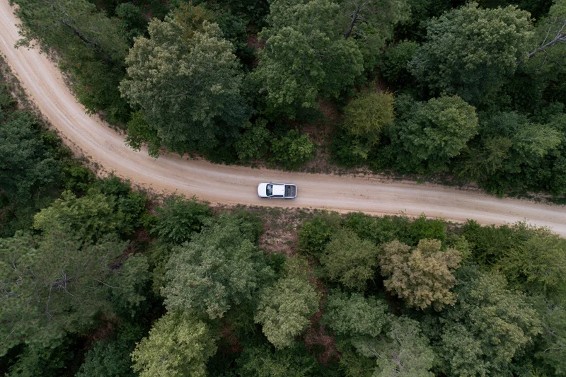 White car driving along winding dirt road through dense forest from aerial top-down view