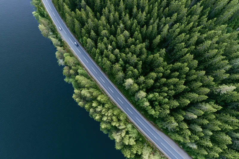 Aerial view of a two-lane road running along a forested shoreline beside a dark lake