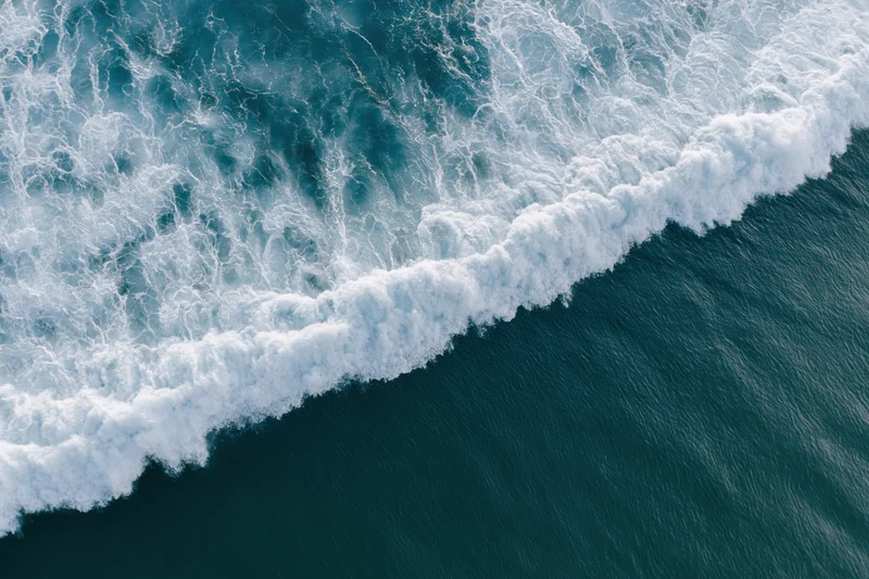 Aerial top-down view of ocean waves breaking in a diagonal line with white foam and deep blue water