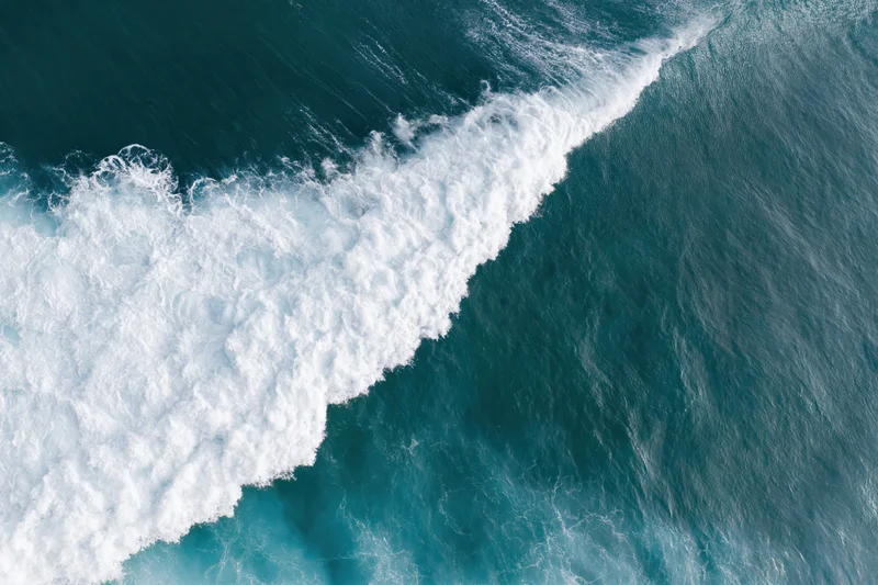 Aerial top-down view of ocean waves breaking in a diagonal line with white foam against blue water