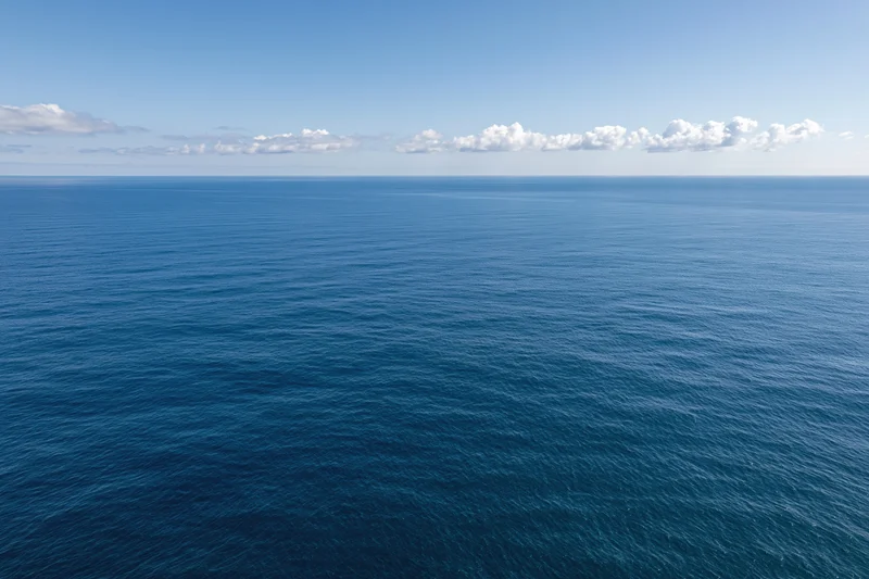 Aerial view of calm open ocean under clear blue sky with scattered clouds along the horizon