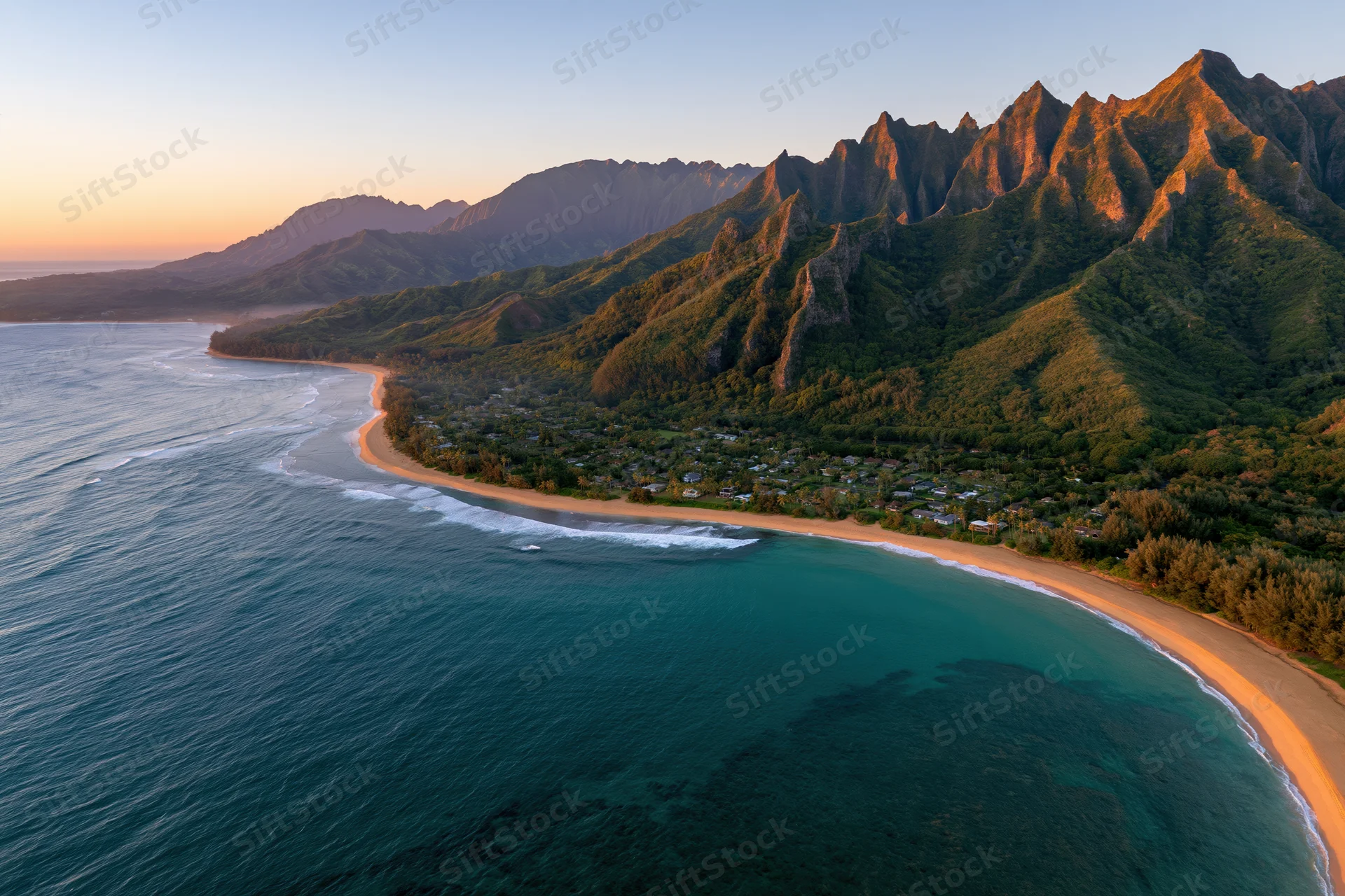 Aerial view of a curved hawaiian beach with golden sand meeting turquoise ocean at sunset beside forested mountains