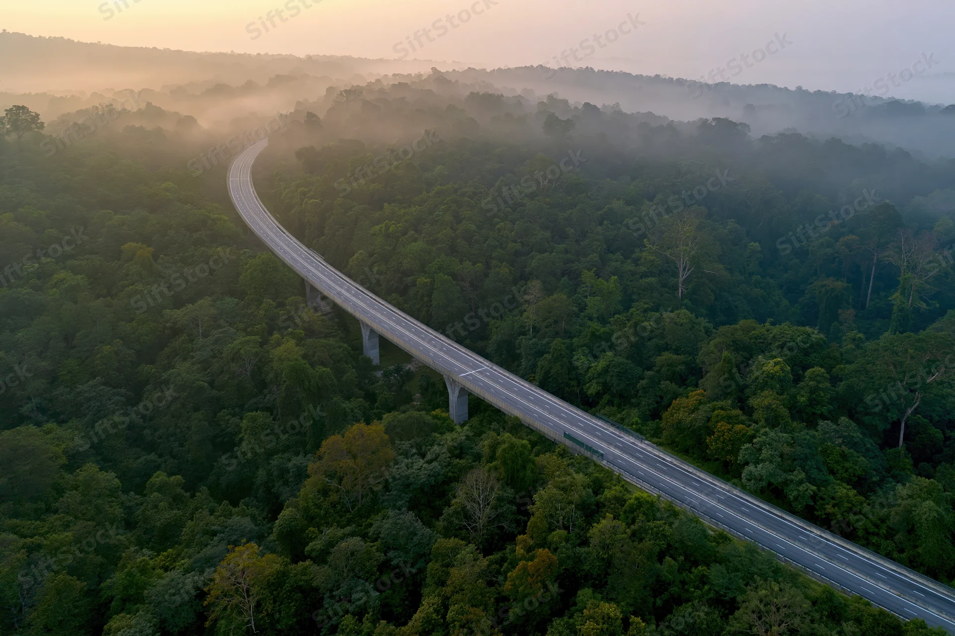 Aerial view of a curved elevated highway viaduct winding through a misty forest at sunrise