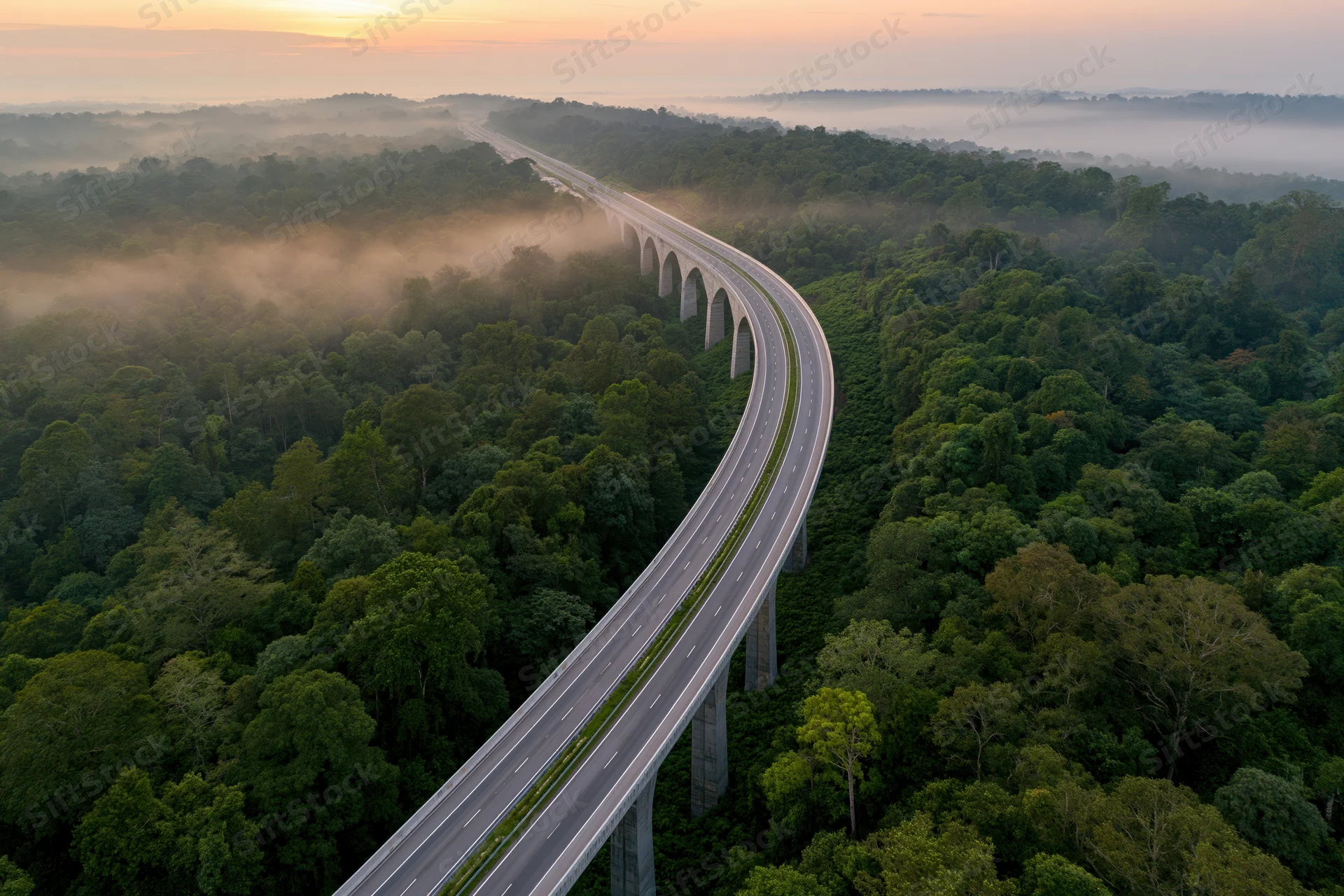 Aerial view of a curved elevated highway viaduct winding through a misty forest at sunrise
