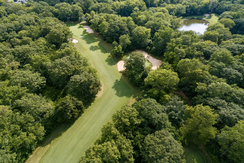 Aerial view of a golf course fairway winding through dense trees with sand traps and a pond nearby
