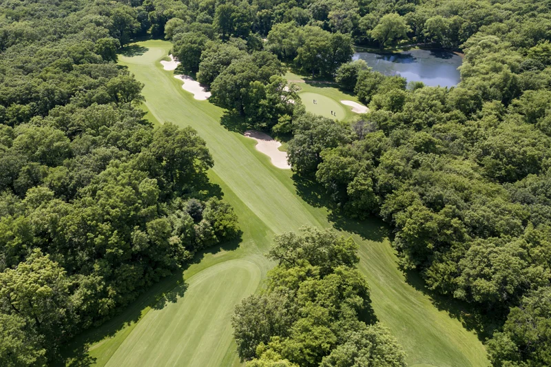 Aerial view of a golf course fairway winding through dense trees with a pond and sand traps visible