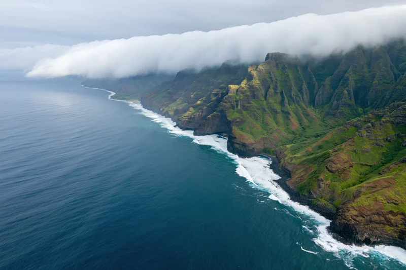 Aerial view of rugged volcanic coastline with steep green cliffs meeting ocean waves under cloudy sky