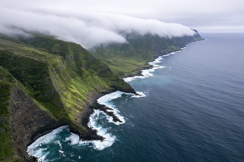 Aerial view of rugged volcanic coastline with steep green cliffs meeting ocean waves under misty clouds