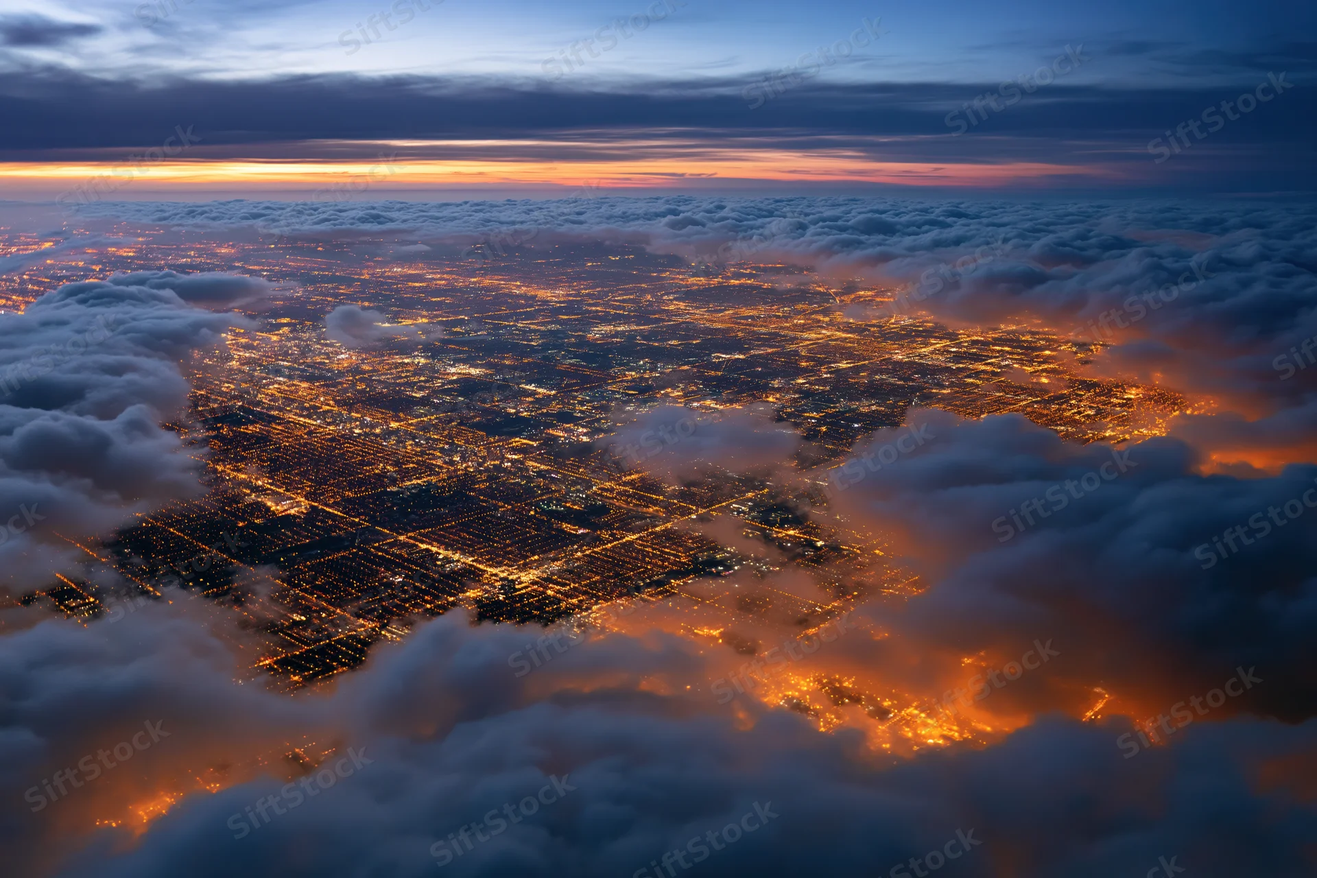 Aerial view of a sprawling city at night from above the clouds, illuminated by streetlights and buildings, seen from an airplane window during twilight
