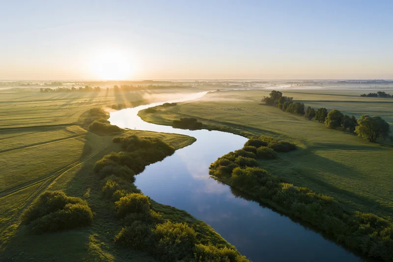 Aerial view of a winding river snaking through flat green fields at sunrise