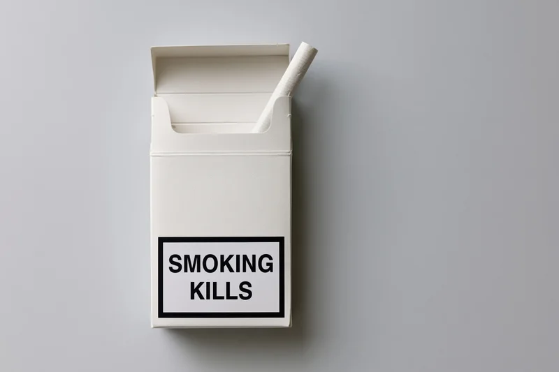 Open white cigarette pack with one cigarette sticking out, warning label reading smoking kills, flat lay on gray surface