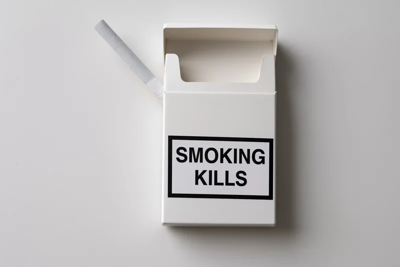 Open white cigarette pack with one cigarette sticking out and smoking kills warning label on plain surface