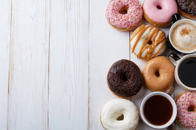 Assorted glazed donuts and ceramic coffee mugs arranged along the right side of a white wooden table in a flat lay top view with copyspace on the left