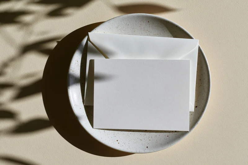 Blank white greeting card and matching envelope on a speckled ceramic plate with leaf shadows