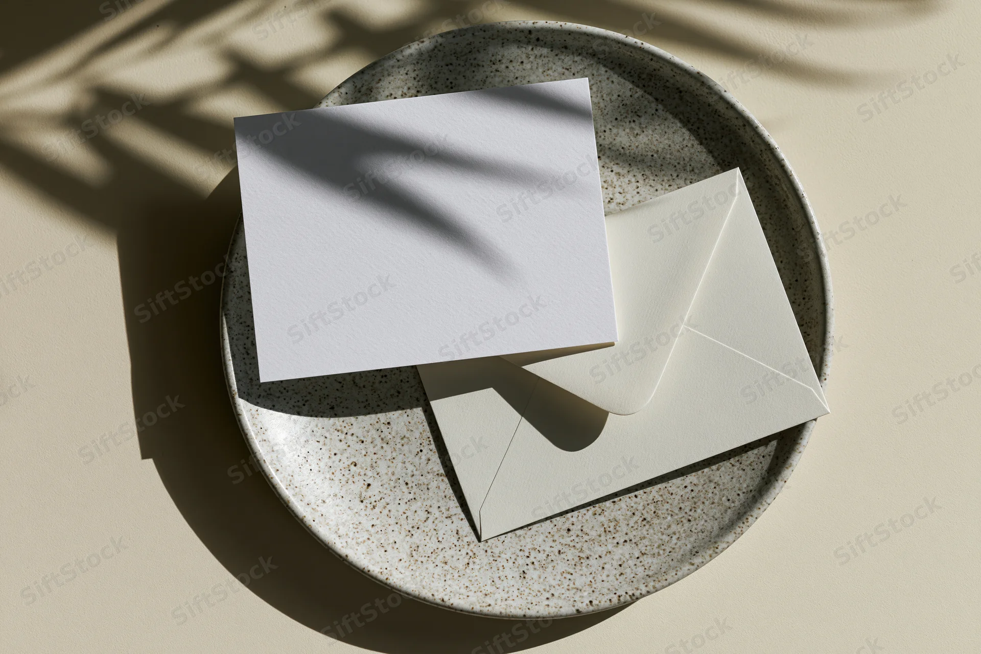 Blank white greeting card and matching envelope on a speckled ceramic plate under natural sunlight with shadows from foliage