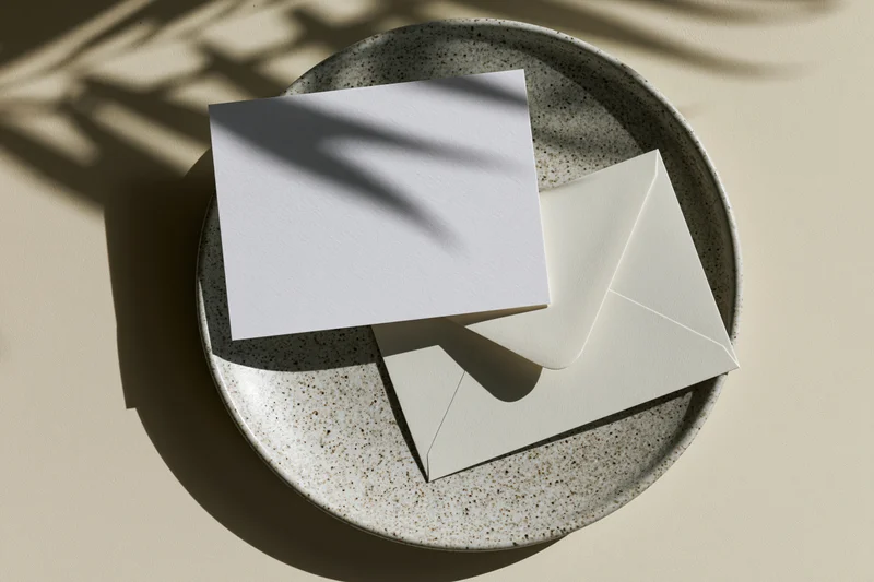 Blank white greeting card and matching envelope on a speckled ceramic plate under natural sunlight with shadows from foliage