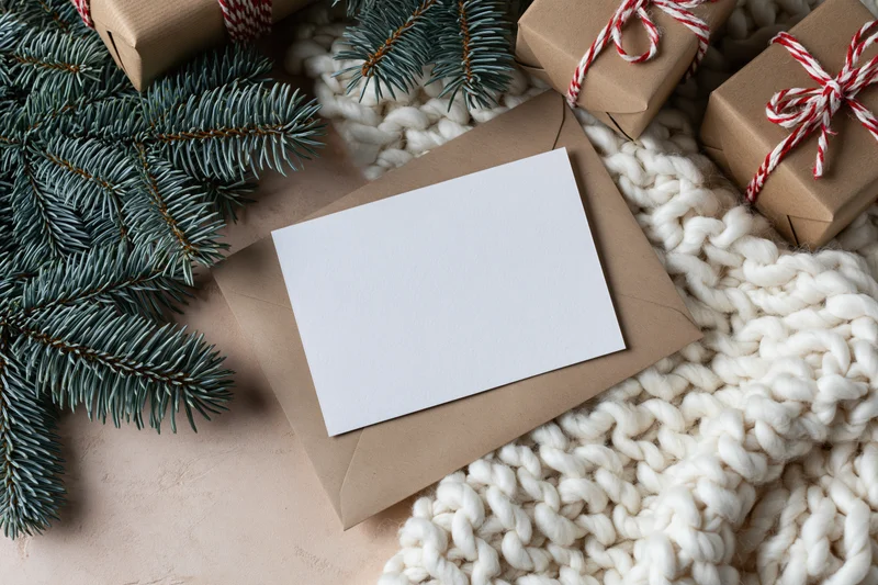 Blank white greeting card on kraft envelope with pine branches and wrapped gifts on knitted wool blanket