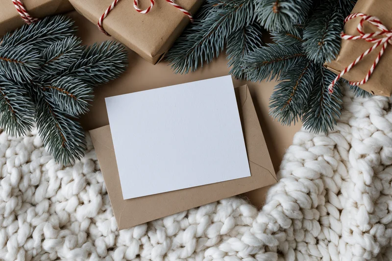 Blank white card on kraft envelope surrounded by pine branches and knit blanket