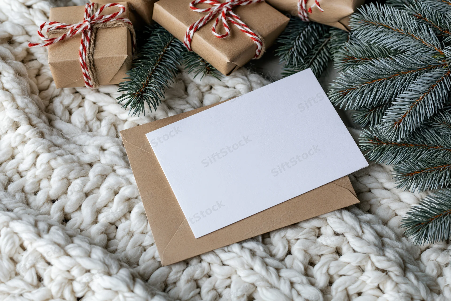 Blank white greeting card on kraft envelope with pine branches and wrapped gifts on knitted blanket