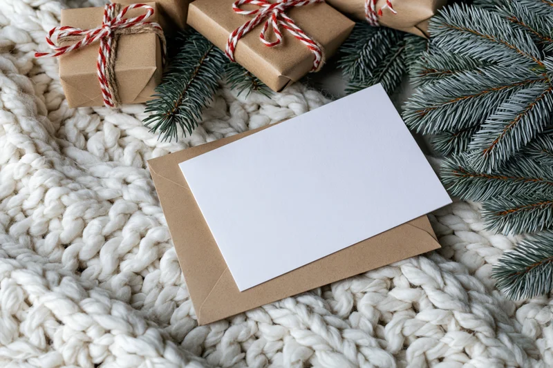 Blank white greeting card on kraft envelope with pine branches and wrapped gifts on knitted blanket