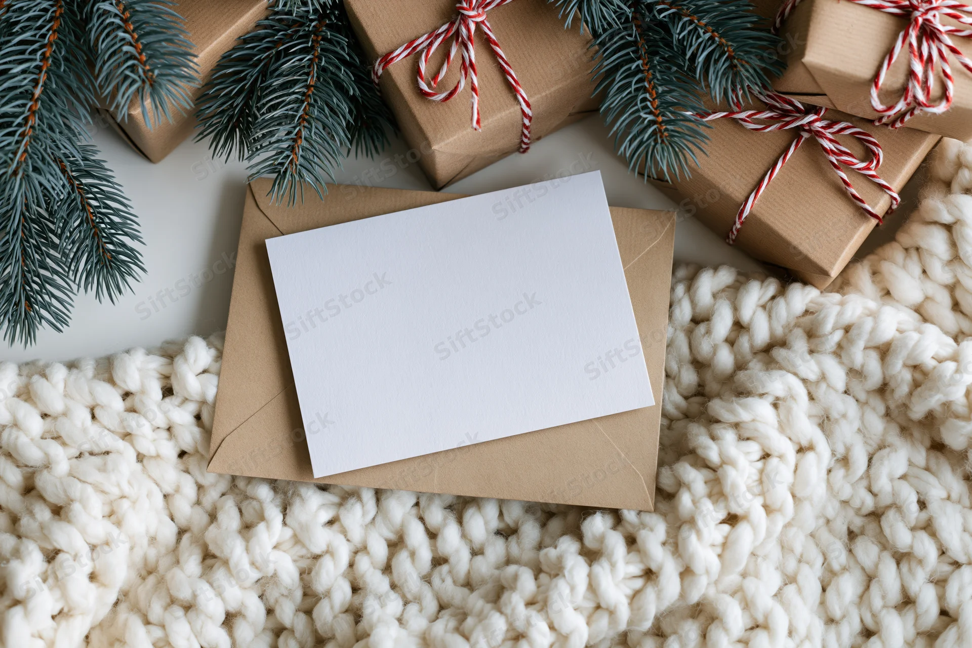 Blank white greeting card on kraft envelope with wrapped gifts and pine branches on knitted wool surface