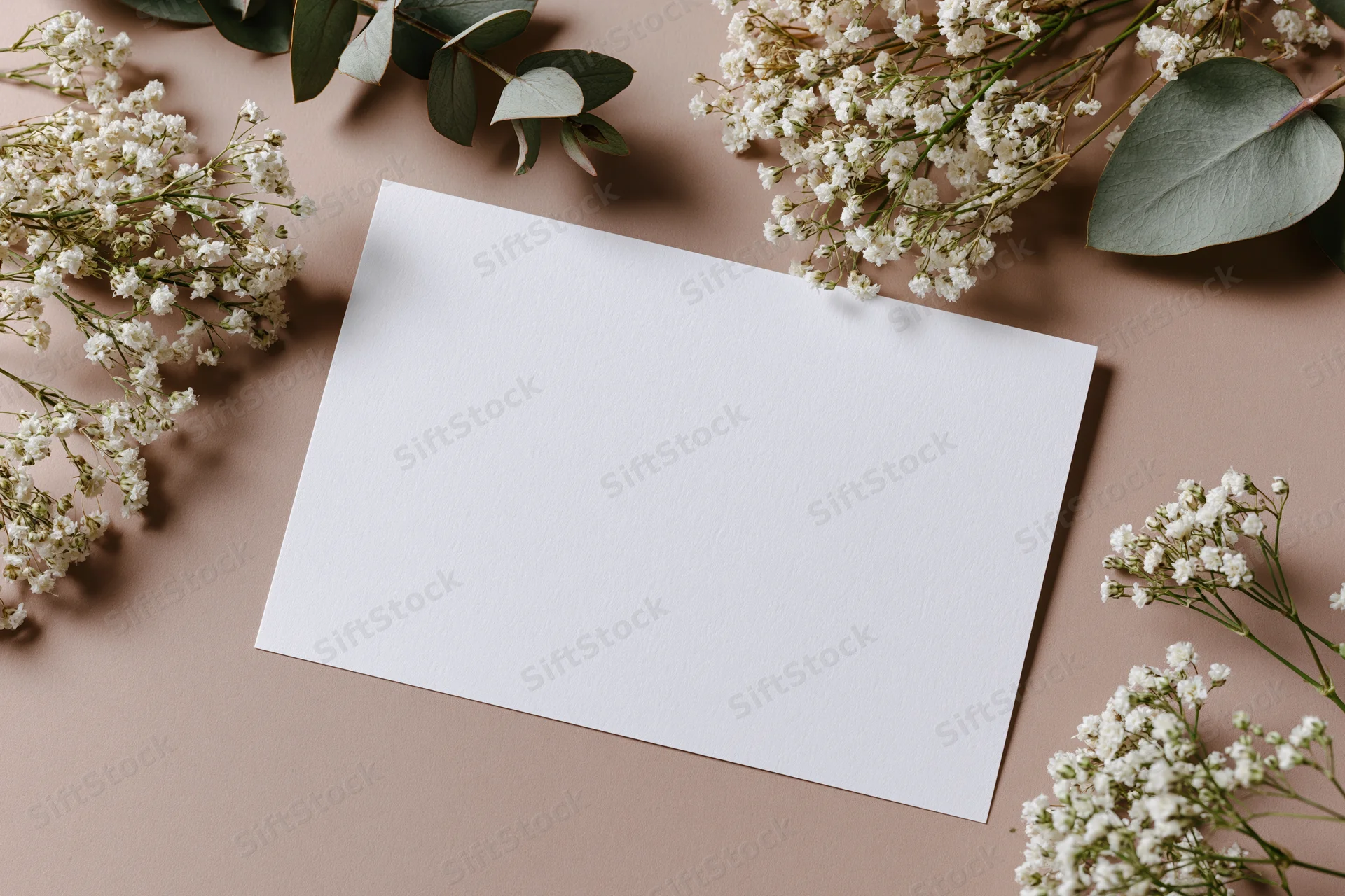 Blank white card on warm taupe surface surrounded by baby's breath and eucalyptus leaves