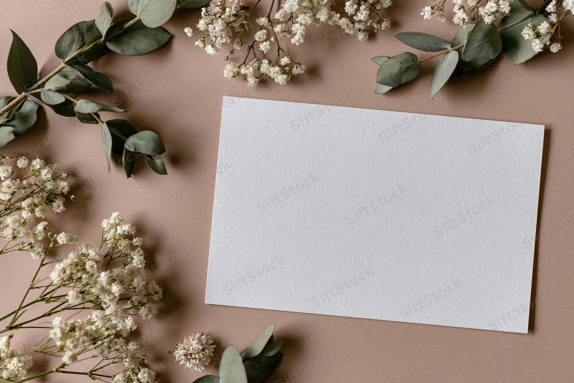 Blank white card on warm taupe surface surrounded by eucalyptus and baby's breath flowers