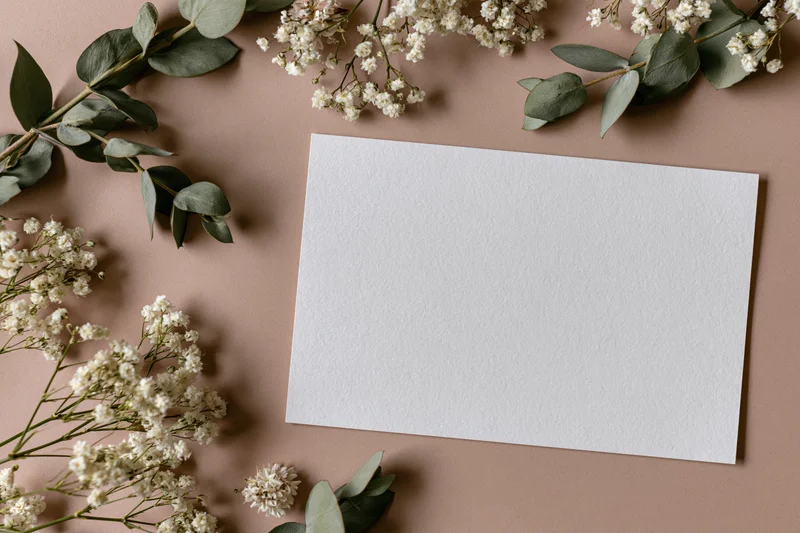 Blank white card on warm taupe surface surrounded by eucalyptus and baby's breath flowers