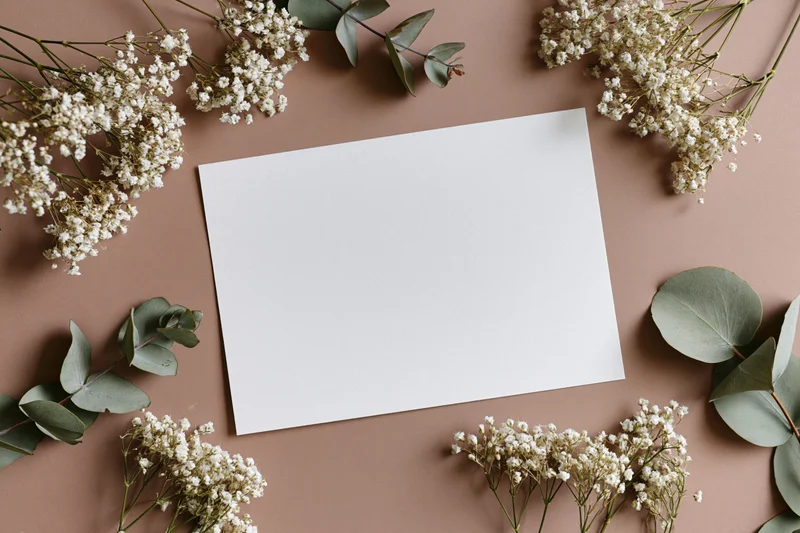 Blank white card on warm taupe surface surrounded by baby's breath and eucalyptus leaves in flat lay arrangement