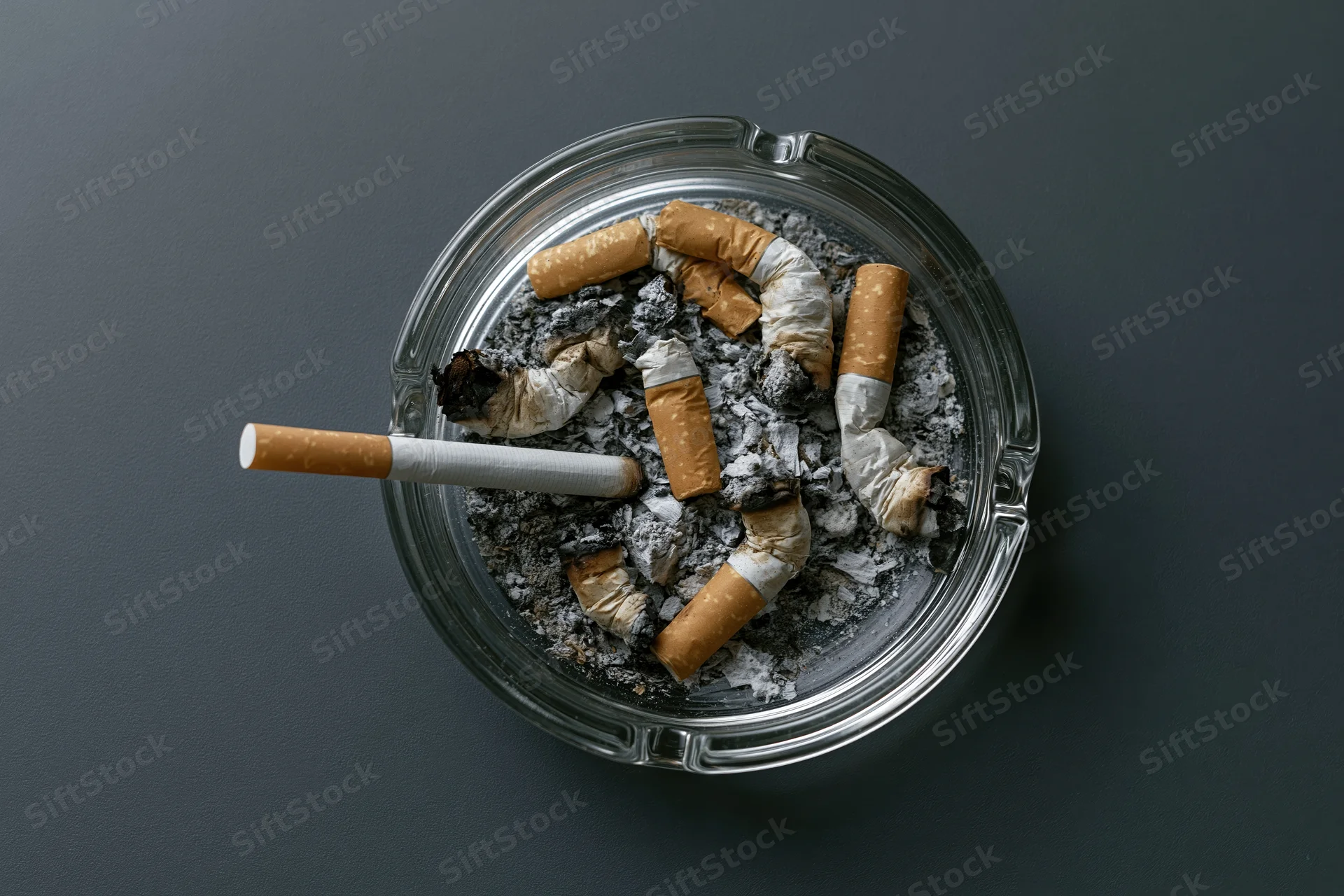 Clear glass ashtray filled with crushed cigarette butts and ash on dark surface