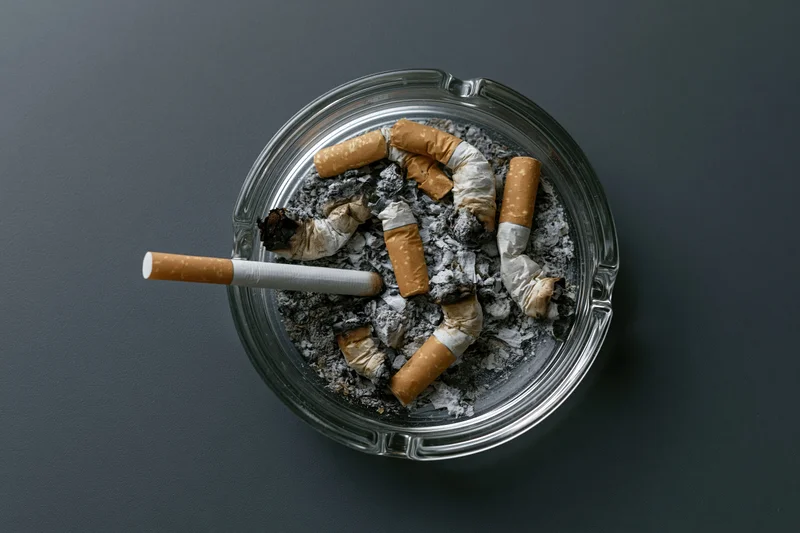 Clear glass ashtray filled with crushed cigarette butts and ash on dark surface