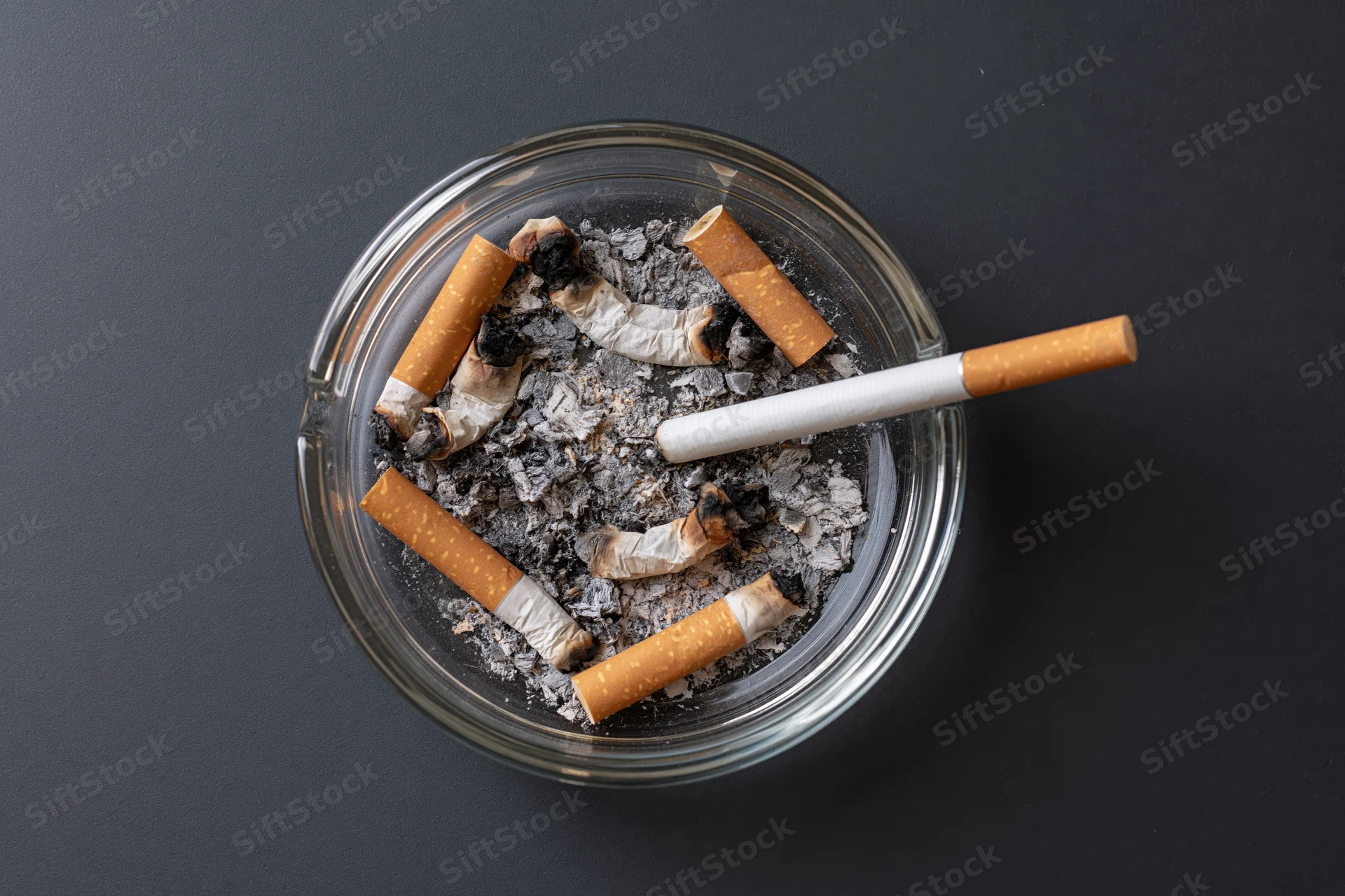 Clear glass ashtray filled with crushed cigarette butts and ash on dark surface