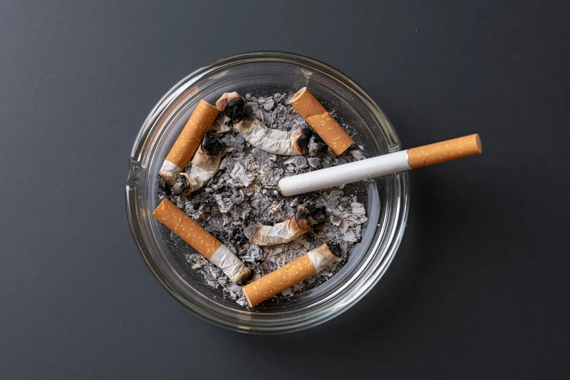 Clear glass ashtray filled with crushed cigarette butts and ash on dark surface