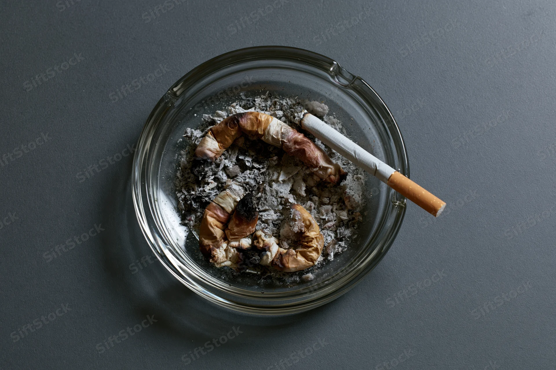 Clear glass ashtray filled with crushed cigarette butts and ash on dark surface