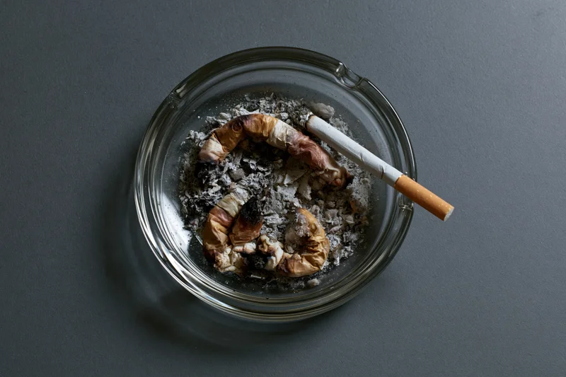 Clear glass ashtray filled with crushed cigarette butts and ash on dark surface