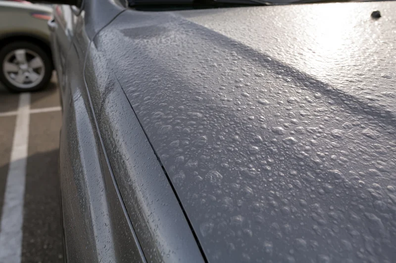 Close-up of dark gray car hood covered in hail damage in a parking lot