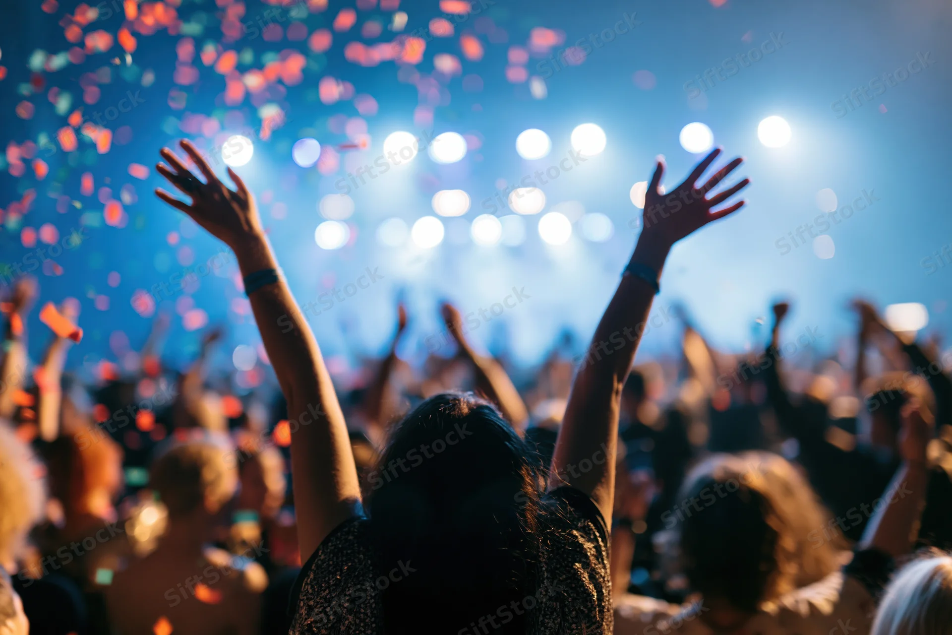 Crowd at concert with arms raised under stage lights and falling confetti