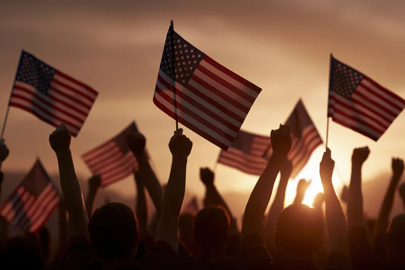 Crowd of people silhouetted from behind with arms raised holding american flags against a sunset sky