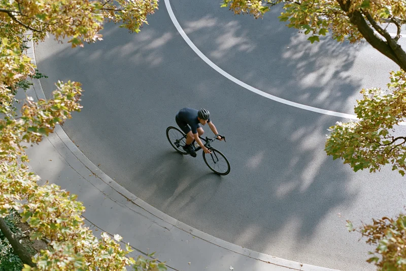 Cyclist riding a road bike along a curved asphalt road framed by autumn trees from above