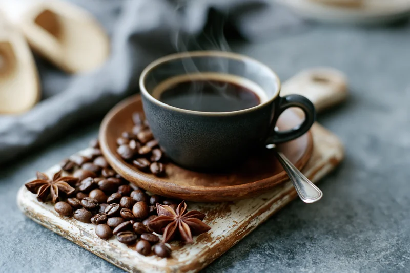 Dark ceramic espresso cup filled with steaming black coffee on a wooden saucer surrounded by coffee beans and star anise on a rustic tray