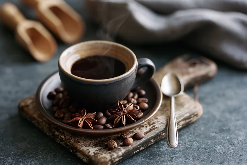 Dark ceramic espresso cup filled with steaming black coffee on a wooden tray with coffee beans and star anise