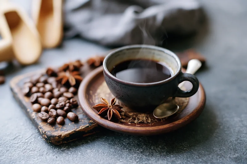 Dark ceramic espresso cup filled with steaming black coffee on a wooden saucer beside coffee beans and star anise on a rustic table