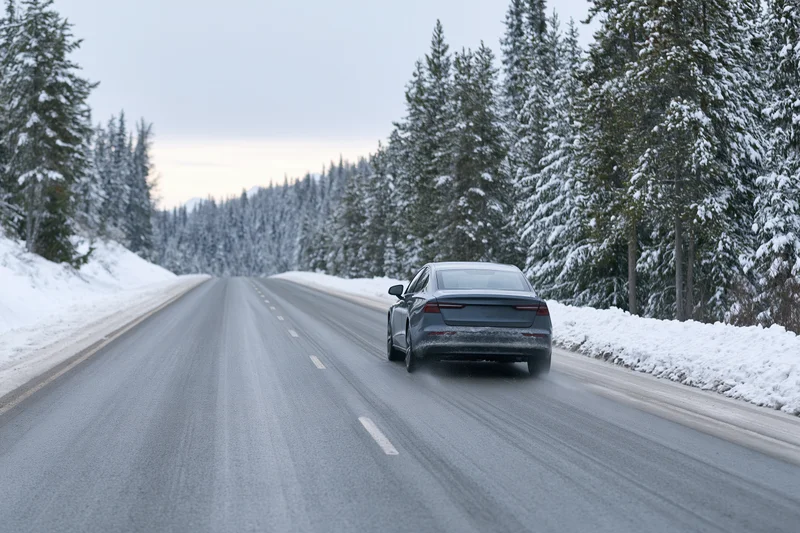 Dark grey electric sedan driving away on snowy road lined with snow-covered pine trees